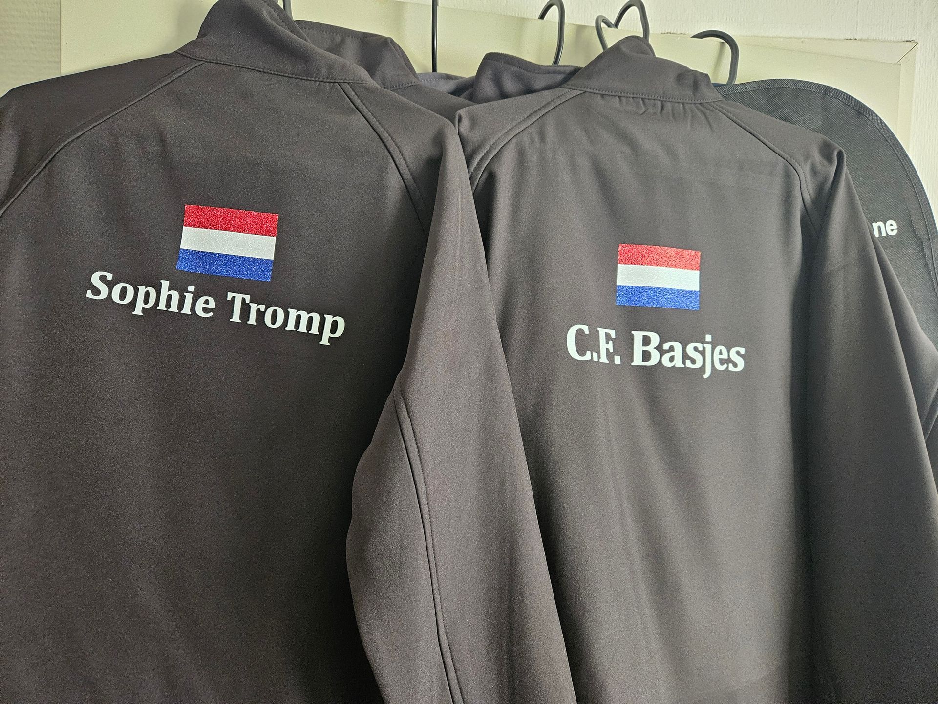 Persoon met zwarte jas met Nederlands vlaglogo, haar in een knot. Buiten met bomen en een veld.