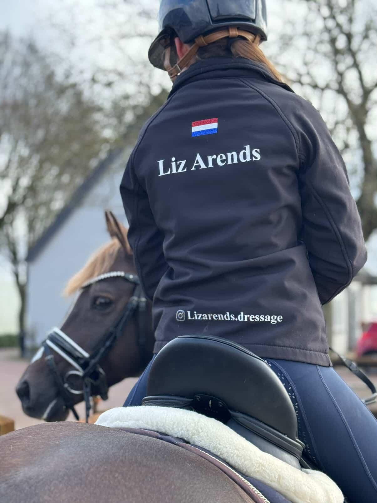 Persoon te paard, gekleed in een zwarte jas met de naam "Liz Arends" en de Nederlandse vlag op de rug.