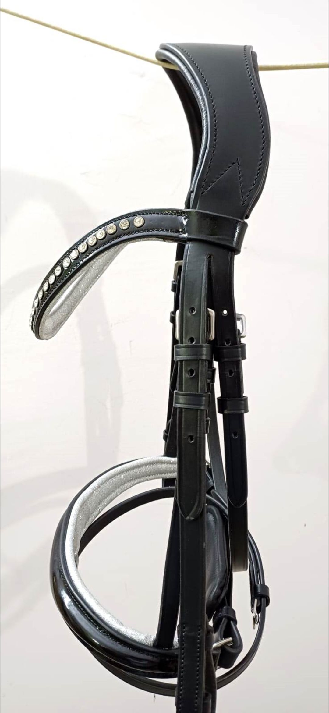 Een zwart leren paardenhoofdstel met witte vulling op de frontriem en neusriem, verticaal hangend tegen een witte muur.