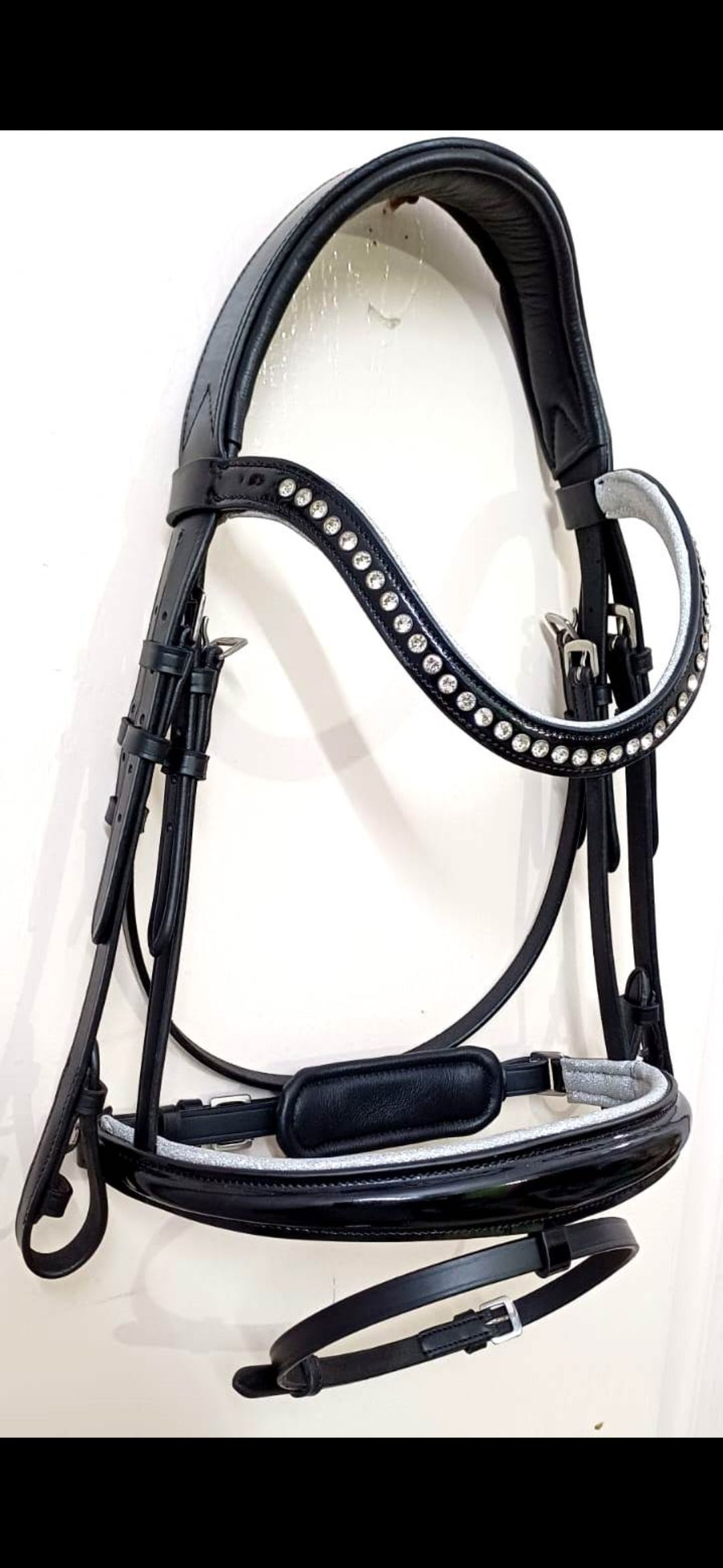 Een zwart leren paardenhoofdstel met witte vulling op de frontriem en neusriem, verticaal hangend tegen een witte muur.