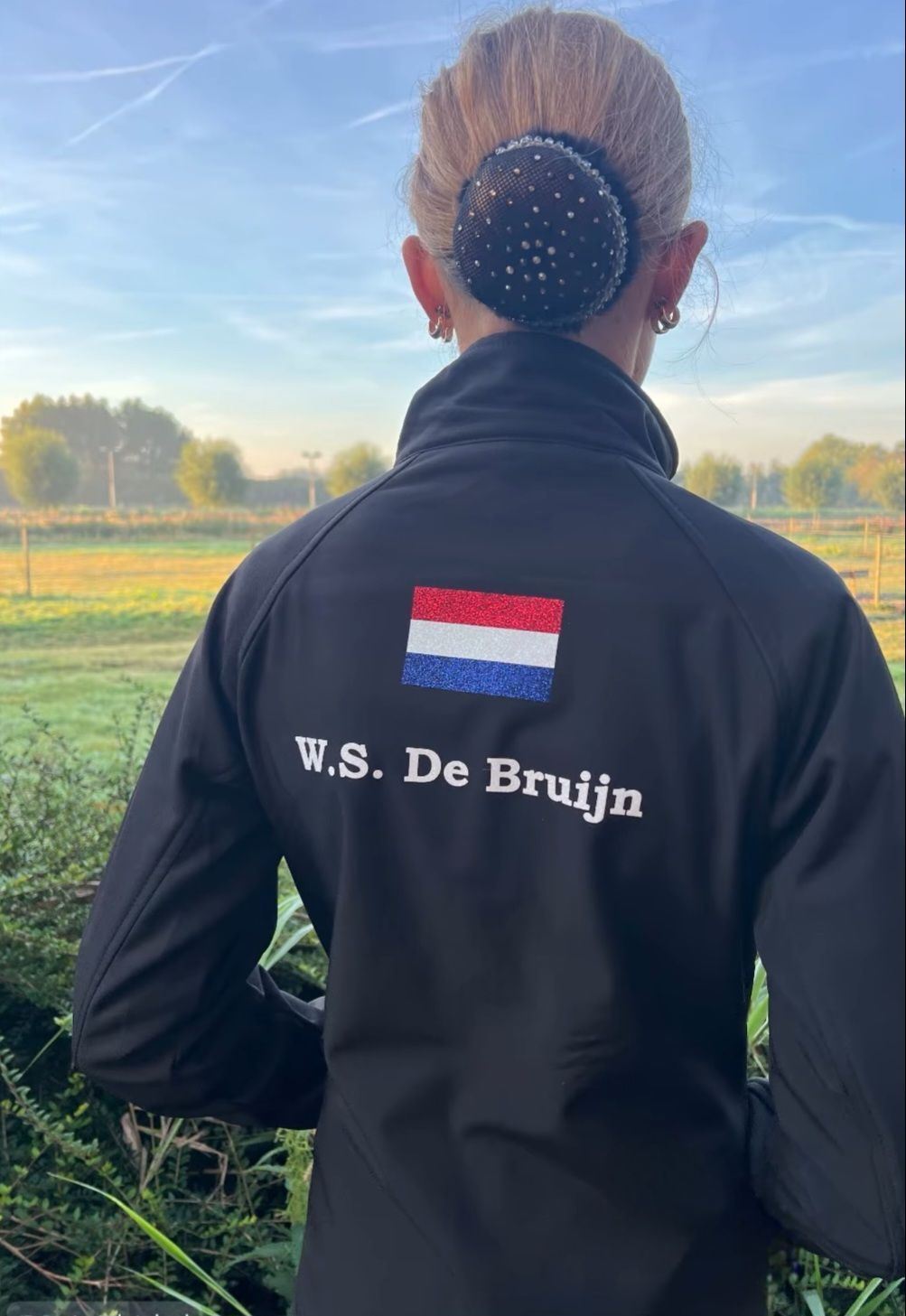 Persoon met zwarte jas met Nederlands vlaglogo, haar in een knot. Buiten met bomen en een veld.