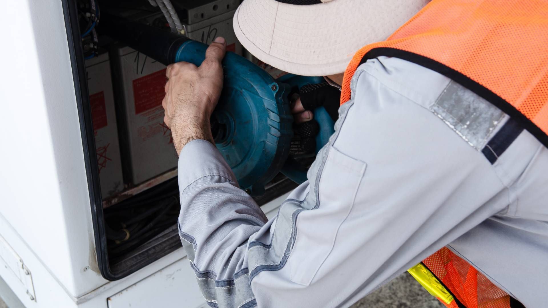 Worker in safety vest using a blue power tool inside a white utility box.