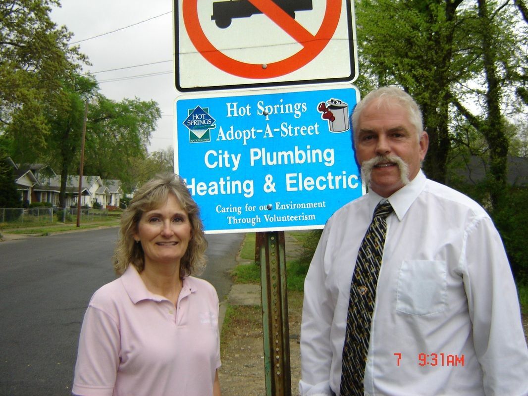 A man and a woman standing in front of a sign that says hot springs adopt-a-street city plumbing heating & electric