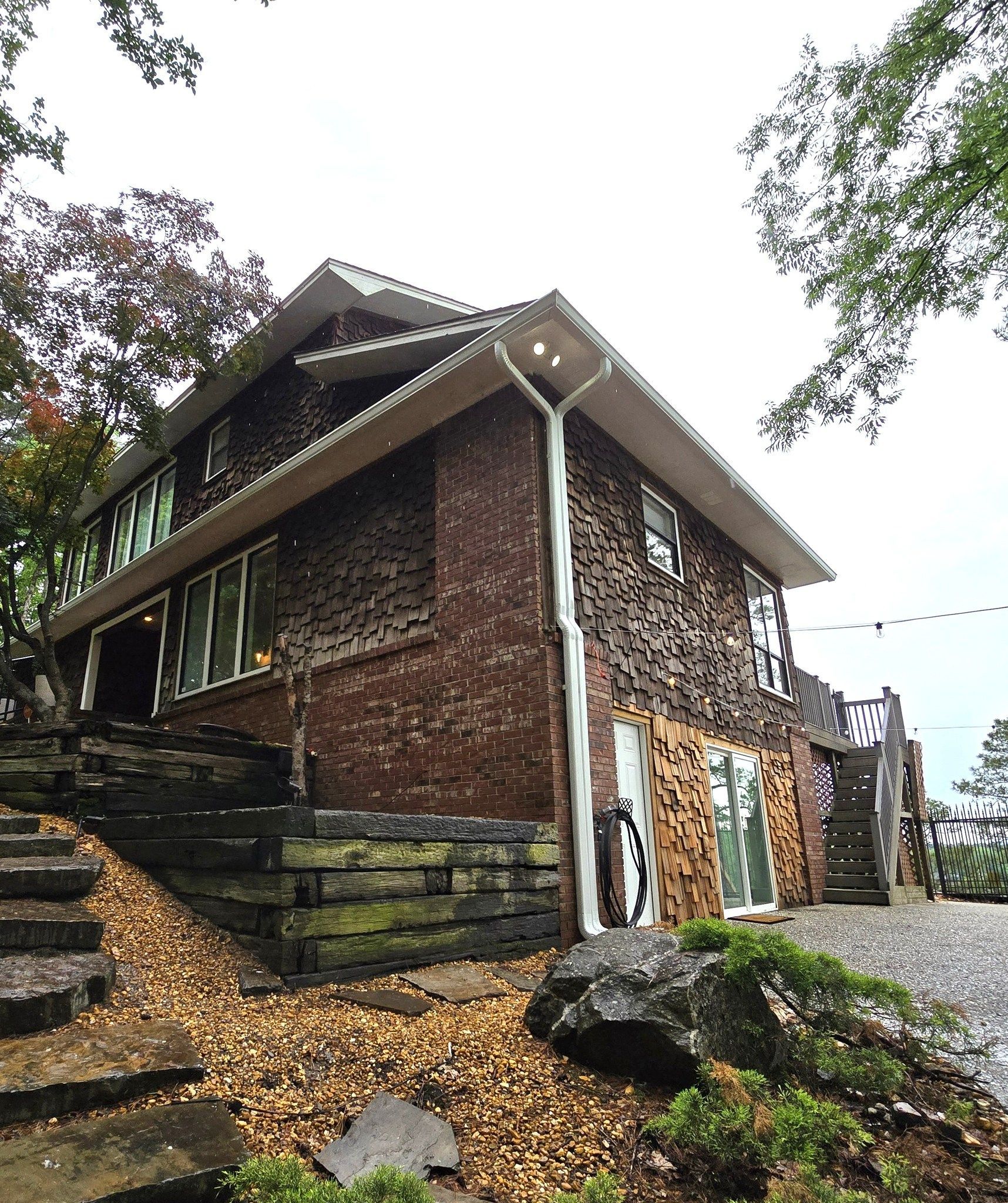 Rustic stone-and-wood house exterior with sloped roof, steps, and trees beside a gravel drive