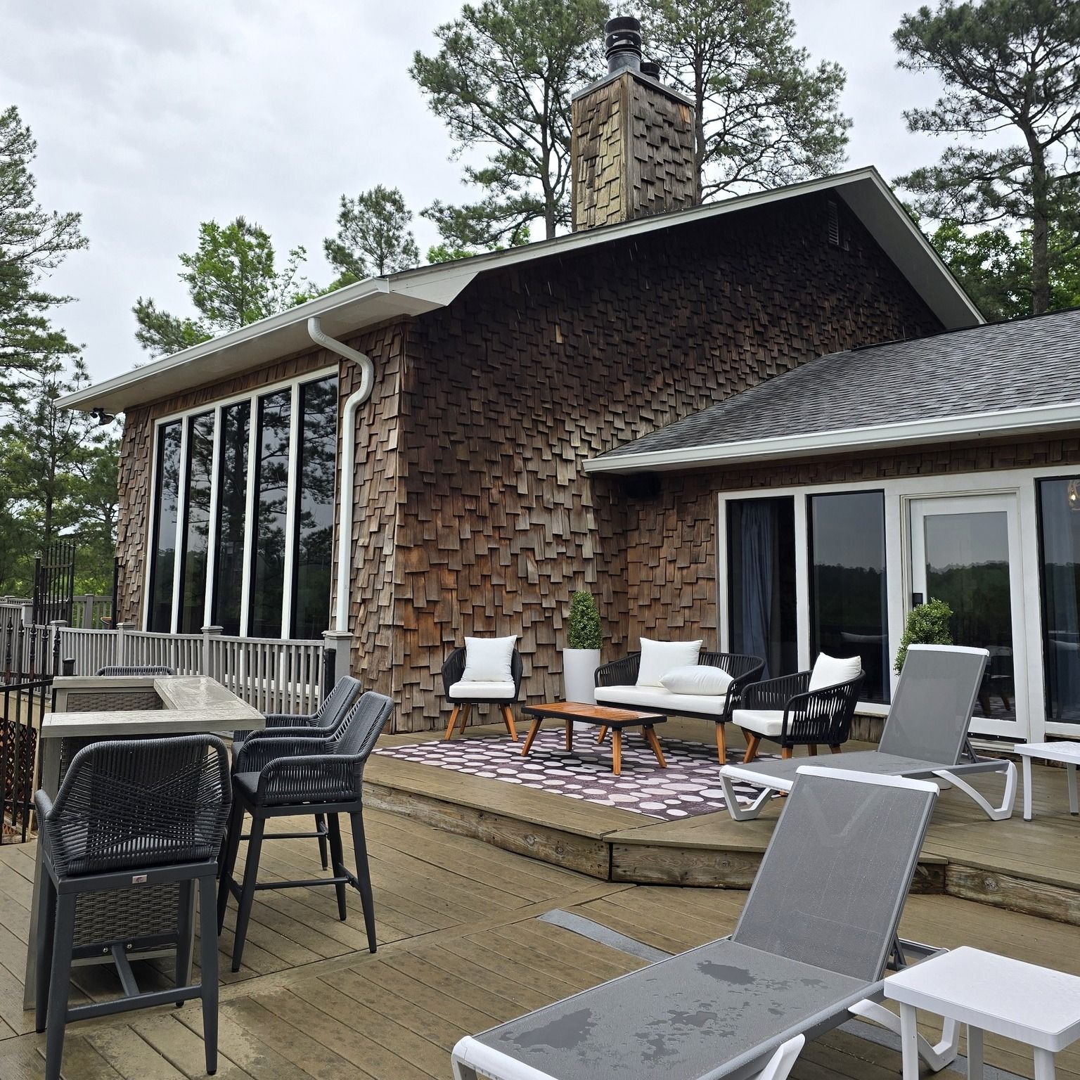 A wooden patio with outdoor seating, including lounge chairs, a fire pit, and a sofa, next to a cabin-style house.