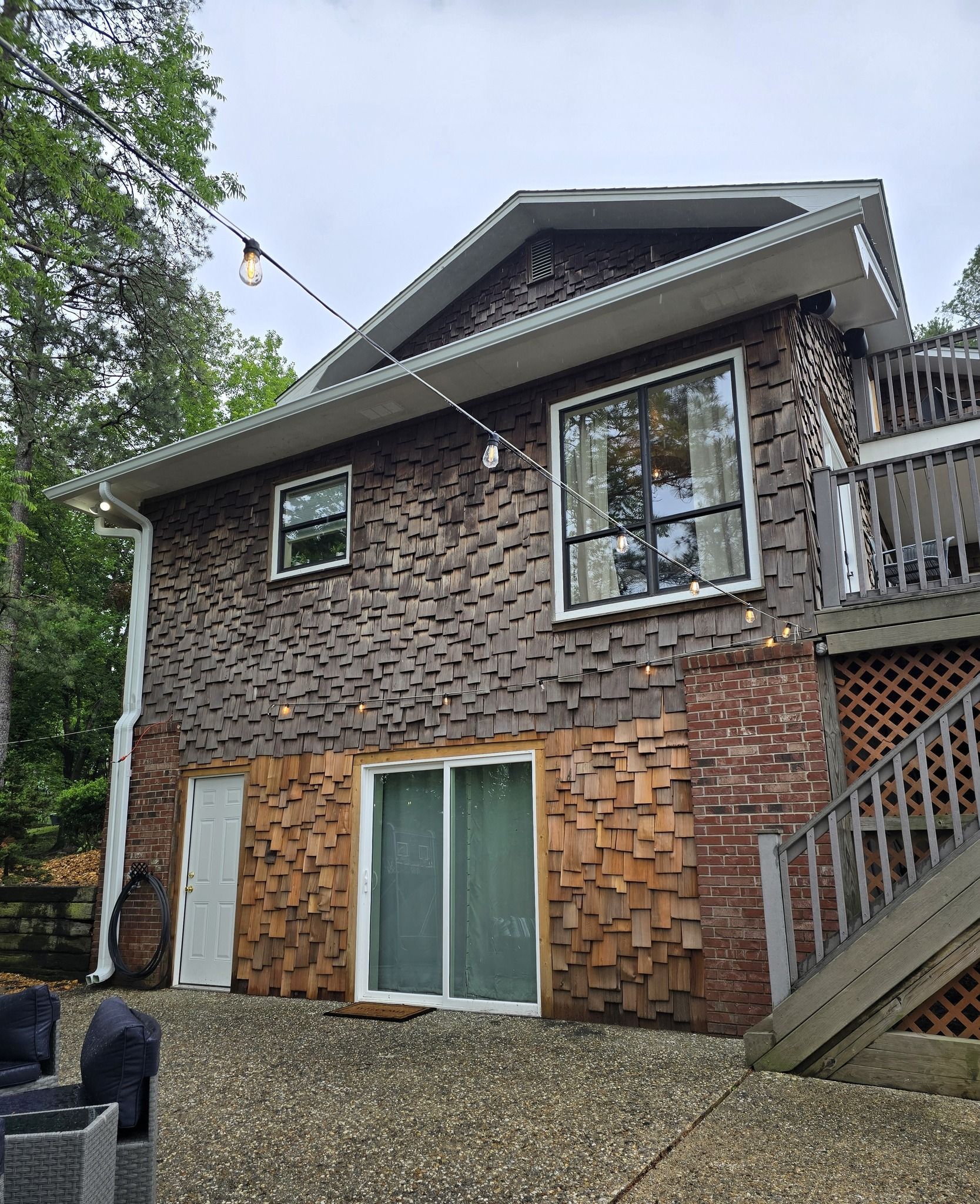 A two-story house with dark textured siding on top and light wooden shingles and brick on the lower level, near a patio.