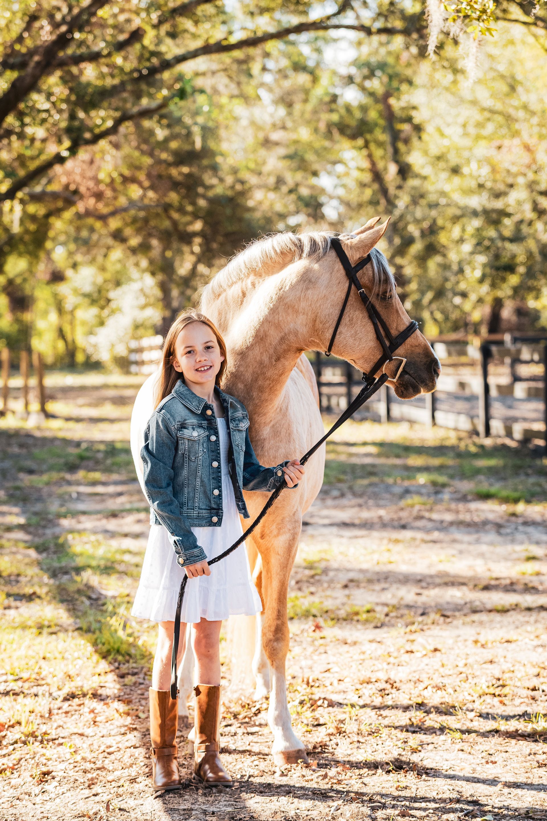 Girl in denim jacket and white dress stands with a tan horse on a path in a sunny field.