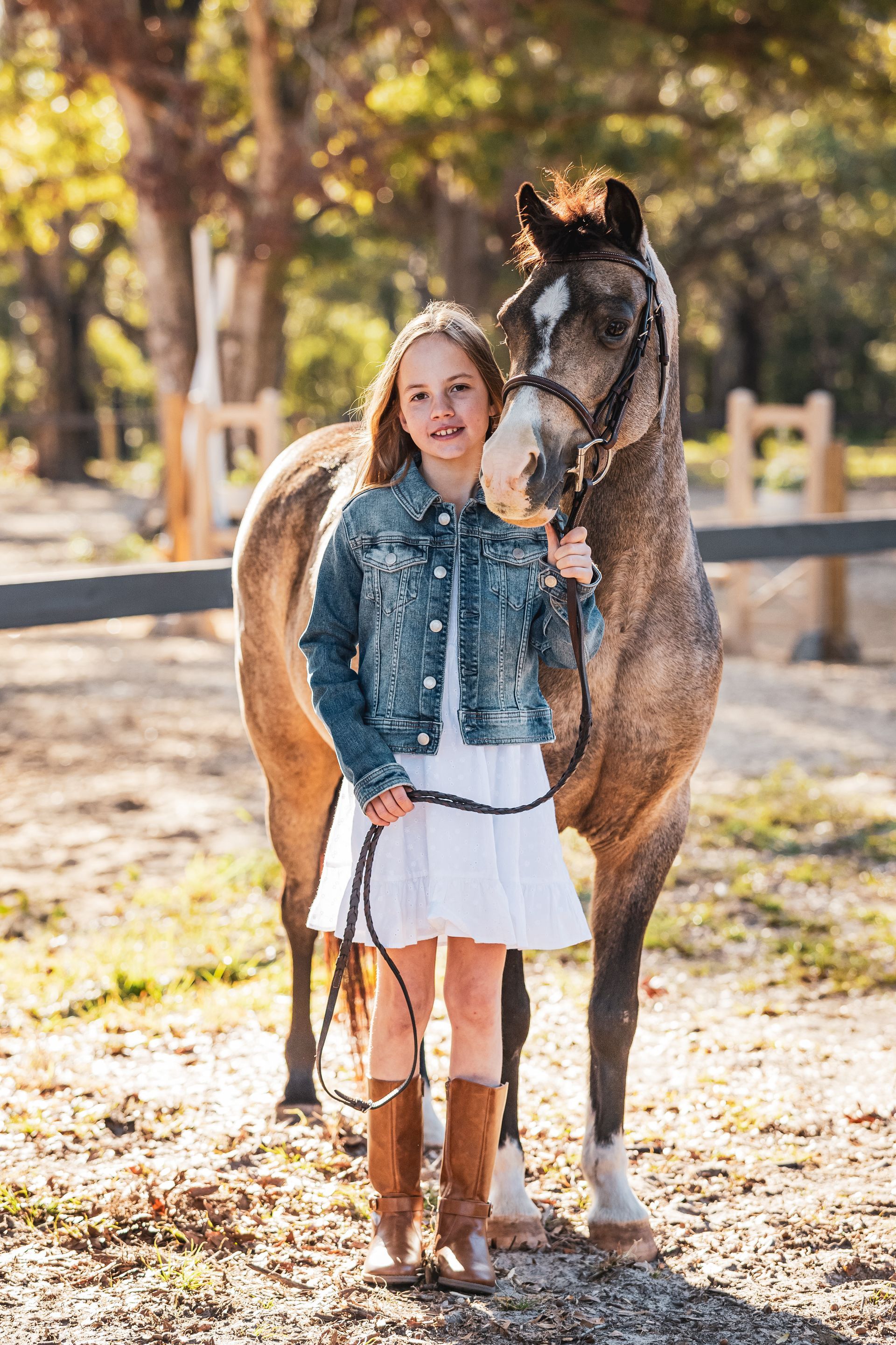 Girl in denim jacket and white dress stands with a dappled horse in a sunny outdoor setting.