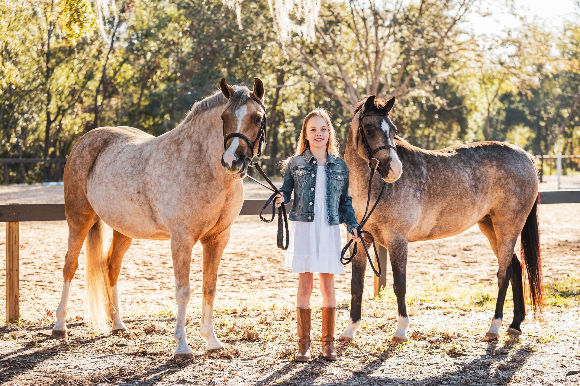 Girl stands between two horses in a sunny, outdoor setting. She wears boots and a denim jacket.