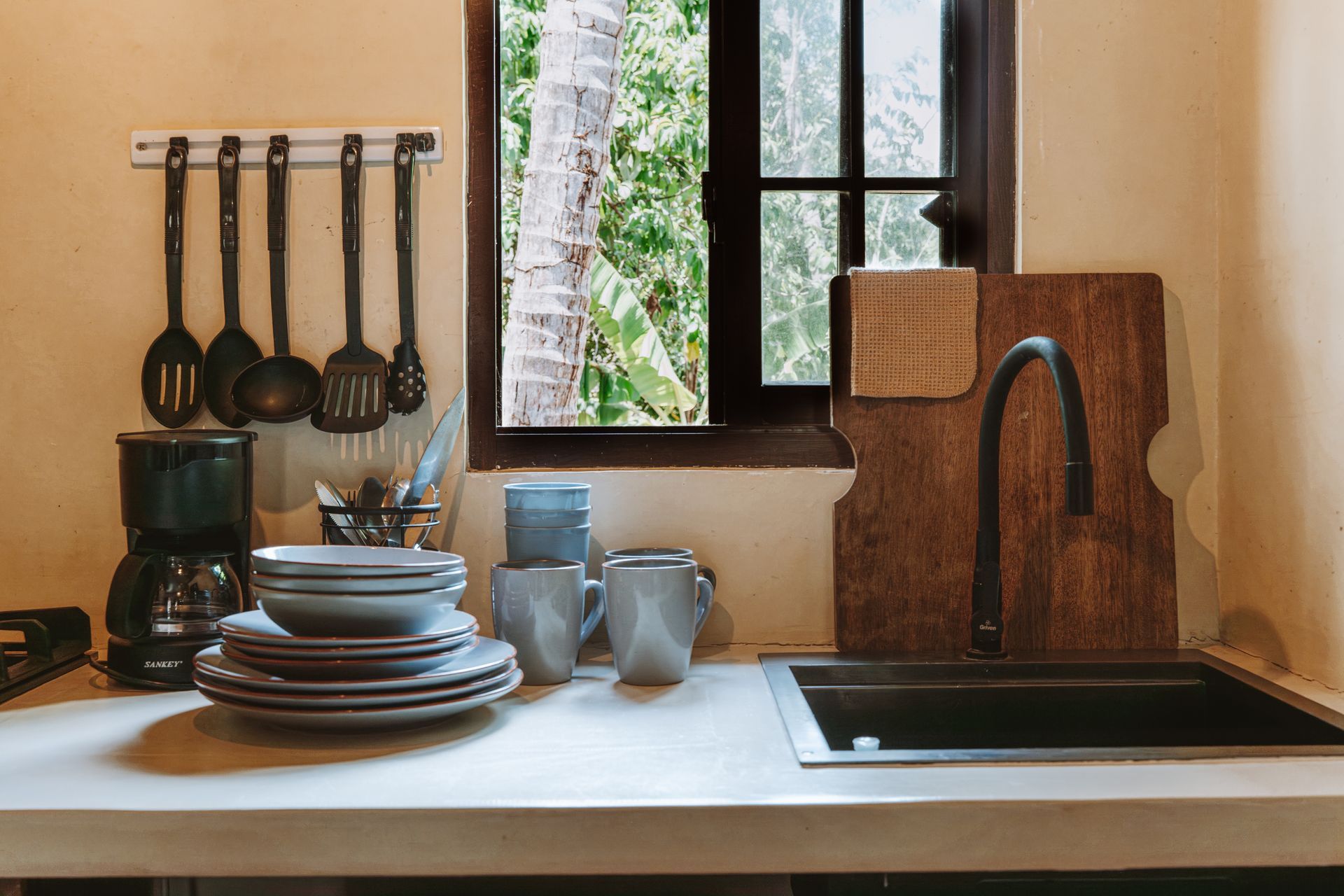 Cozy kitchen sink area with hanging utensils, stacked dishes, mugs, and a window above the counter