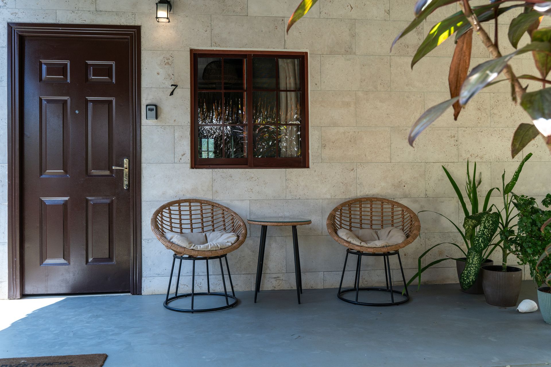 Front porch with dark brown door, window, two wicker chairs, small table, and potted plants.