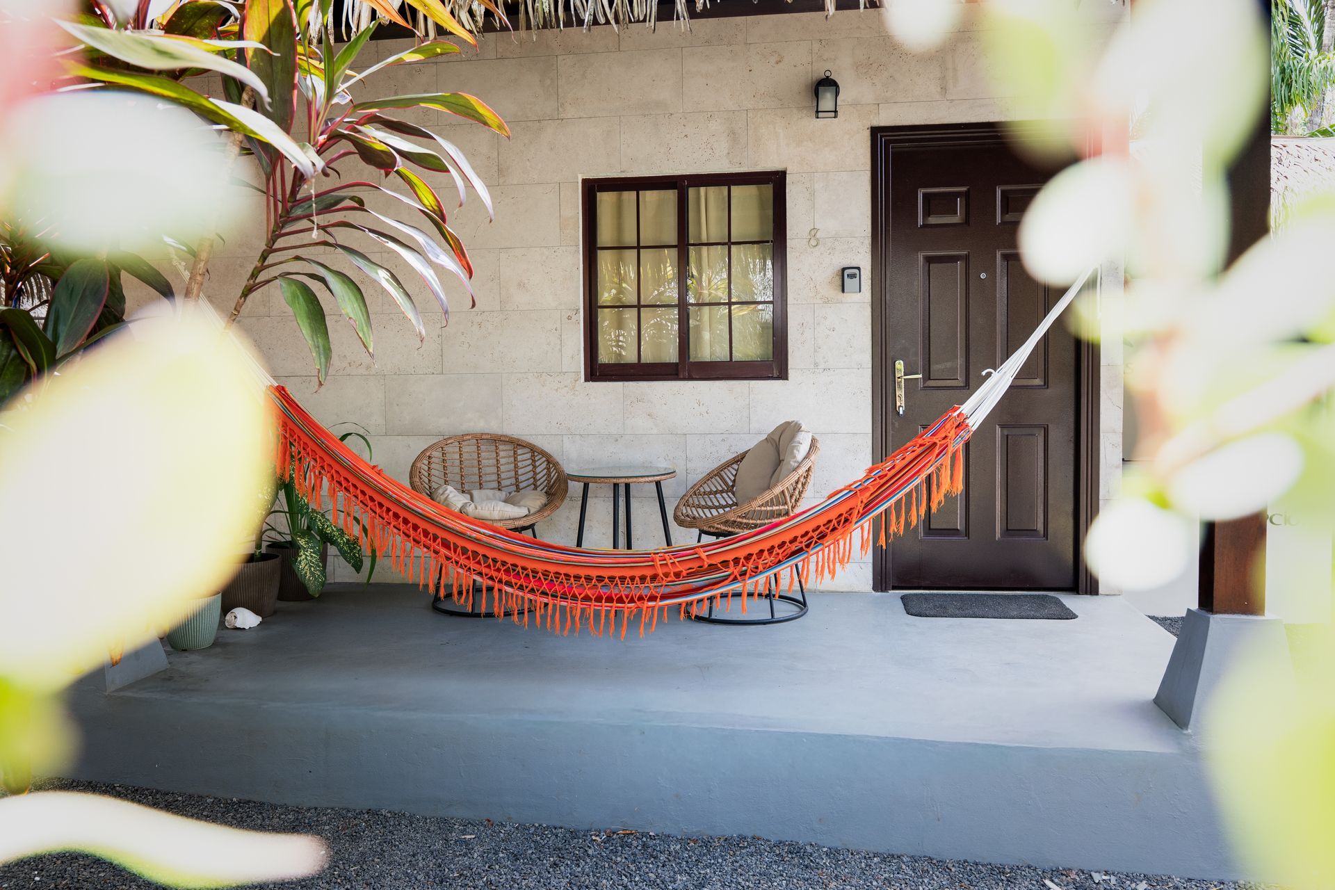 Orange hammock on a porch between chairs, with a door, window, and potted plants.