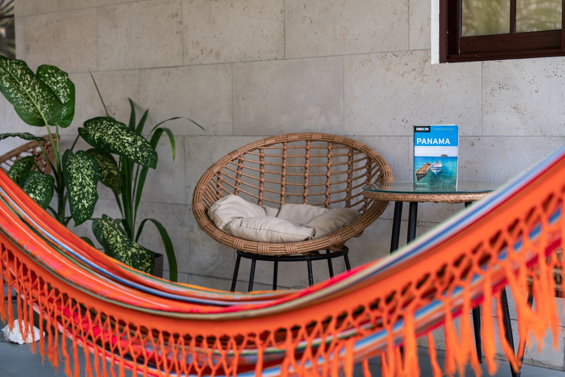 Orange hammock framing a wicker chair with cushion, potted plant, and small table on a stone patio