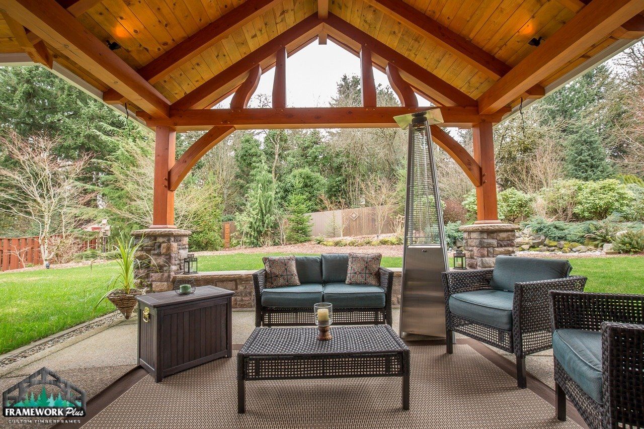 A patio with a couch , chairs , table and heater under a wooden gazebo.