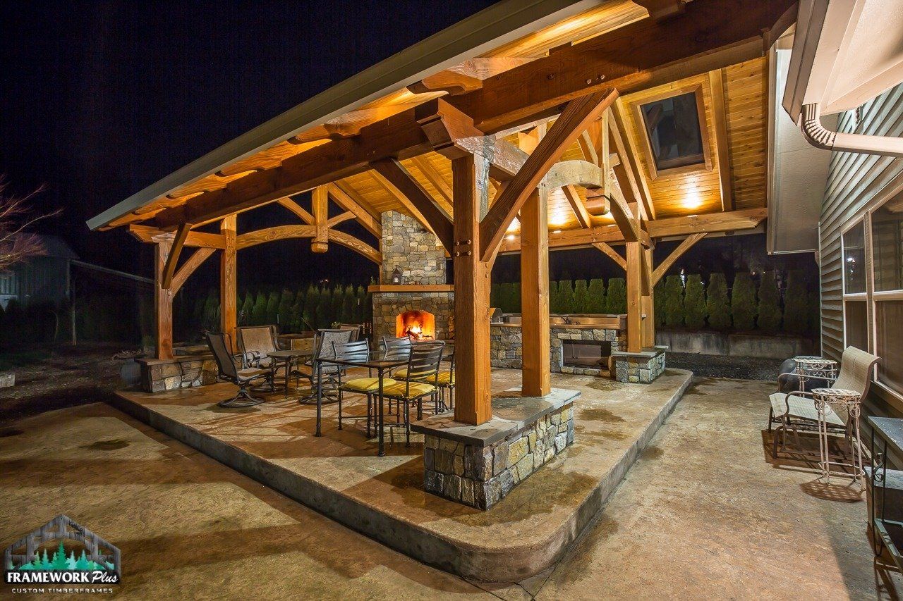 A wooden gazebo with a fireplace and chairs underneath it at night.