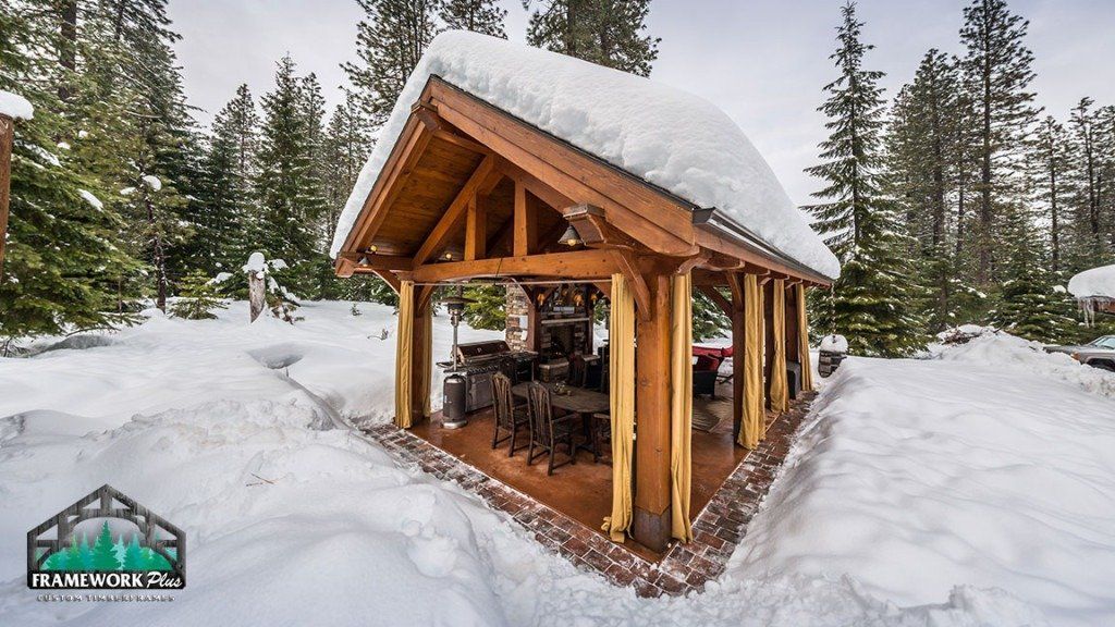 A wooden pavilion with snow on the roof is surrounded by snow covered trees