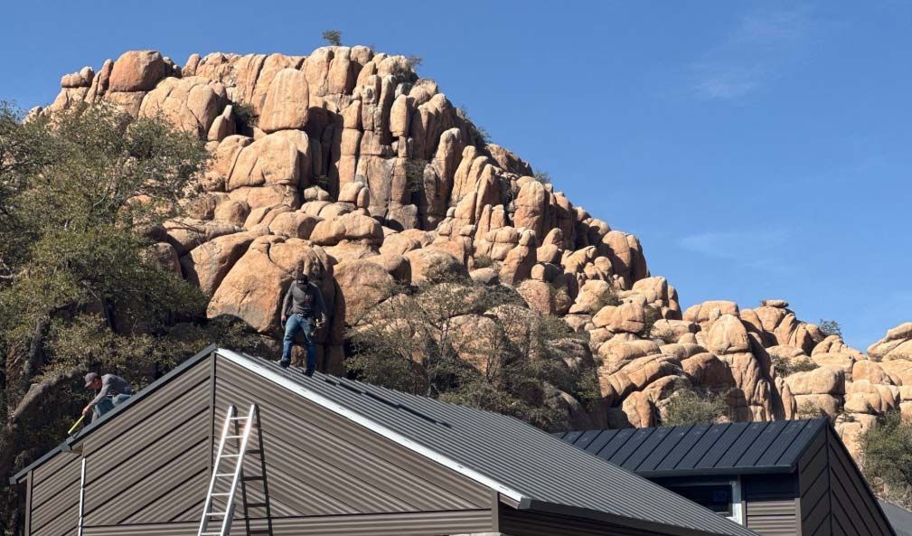Two people on a corrugated metal roof; ladder present. Granite mountain in background, blue sky.