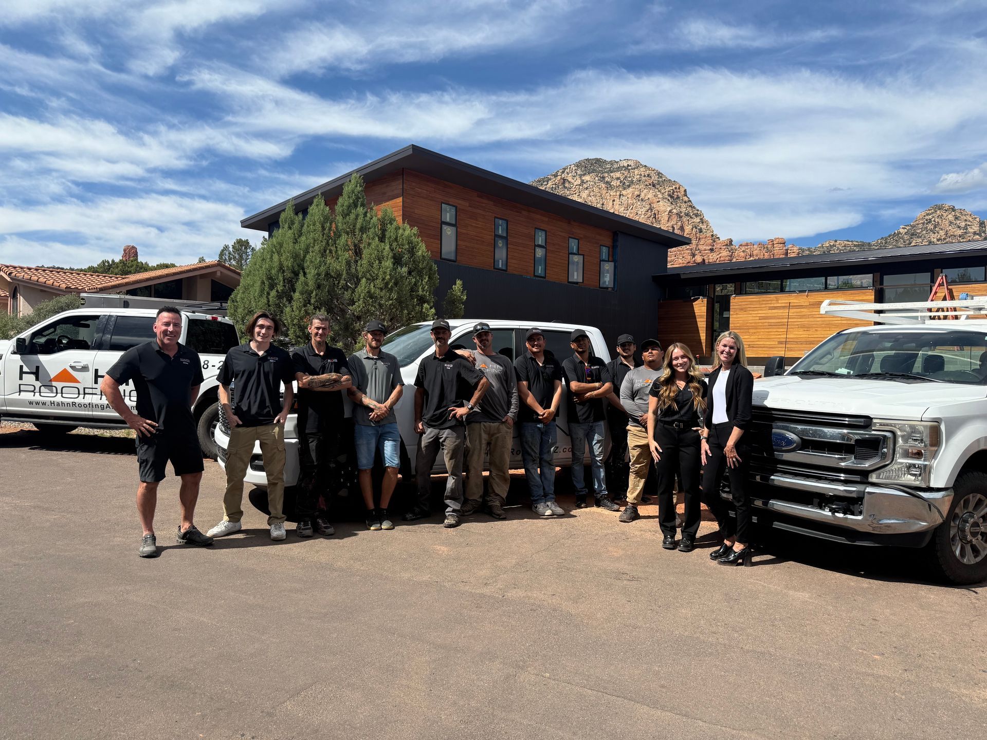 Group of people posing in front of a modern building, flanked by work trucks, on a sunny day.