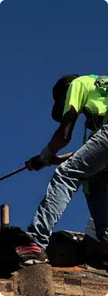 Roofer in safety gear on a roof against a blue sky, using a tool.