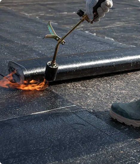 Person using a torch to seal black roofing material on a flat surface.