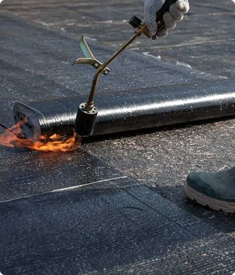 Person using a torch to seal black roofing material on a flat surface.