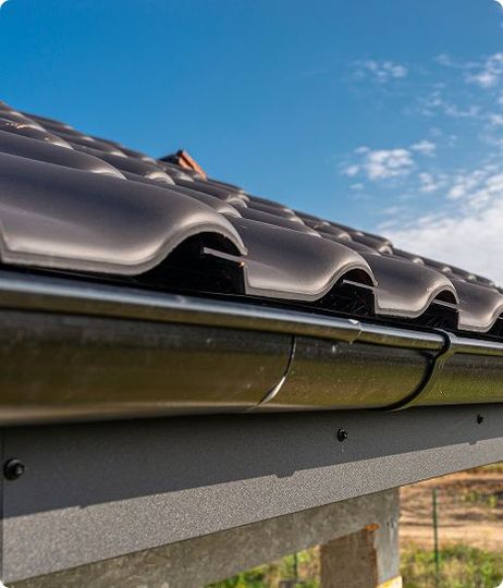 Dark gray roof with curved tiles and black gutter against a blue sky.