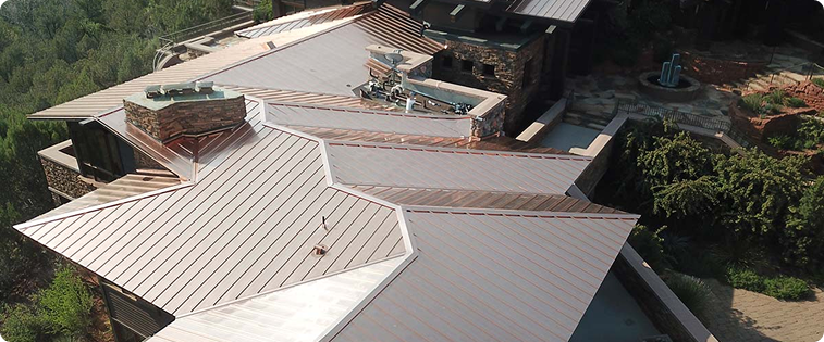 Aerial view of a modern house with a complex, silver metal roof and stone walls, surrounded by trees.