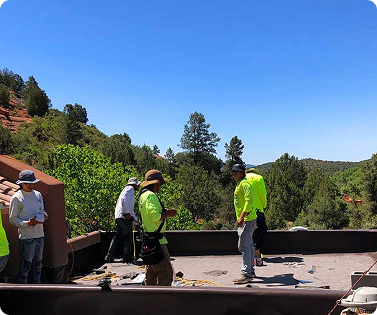Construction workers on a rooftop under a blue sky, surveying the site. Green trees and hillside are in the background.