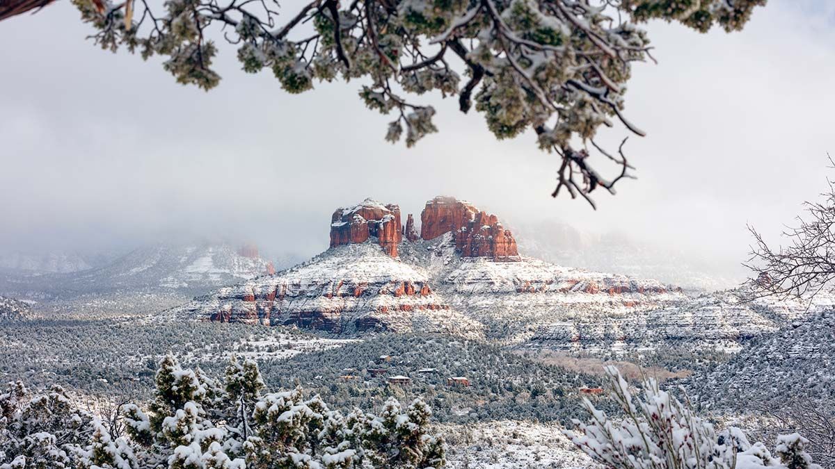Snowy red rock formations in Sedona, Arizona, framed by snow-covered trees and a cloudy sky.
