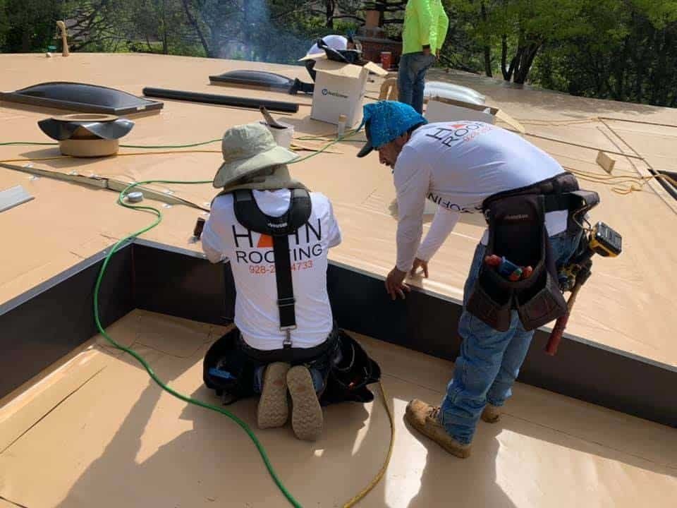 Construction workers installing roofing on a flat roof, outdoors. One kneels, the other points.