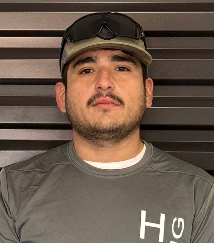 Man wearing a cap and sunglasses, looking at the camera. He has a mustache and a gray shirt with a logo.