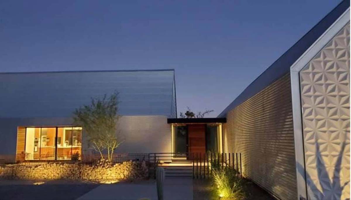 Modern house exterior at dusk, illuminated facade, textured wall, wooden door, pathway, landscaping.