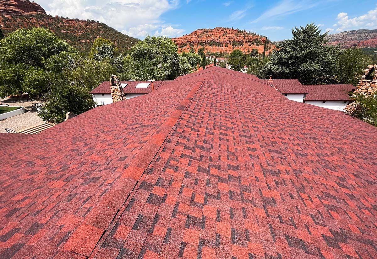 Red-shingled roof with mountain backdrop and trees under blue sky.