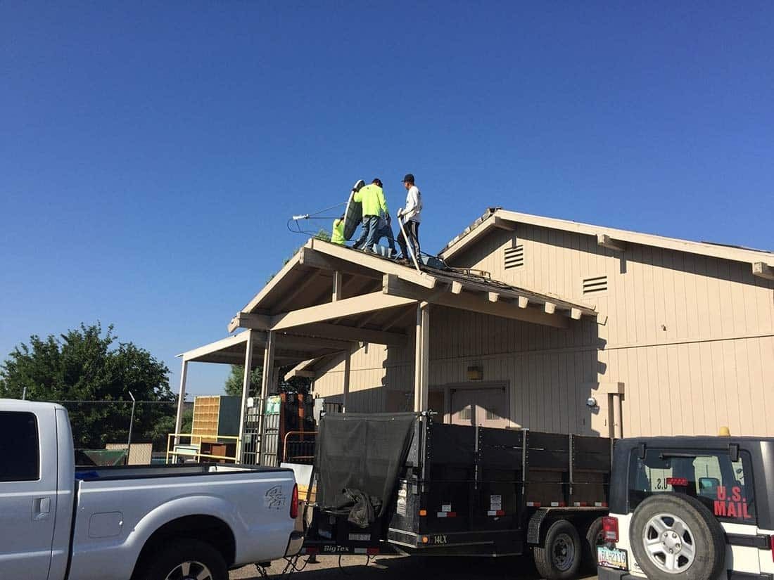 Workers on a roof removing something. White building with a covered porch. A white truck and trailer are in front.