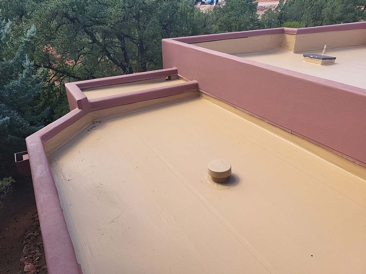Flat roof with light tan surface and surrounding walls, a small vent, and trees in the background.