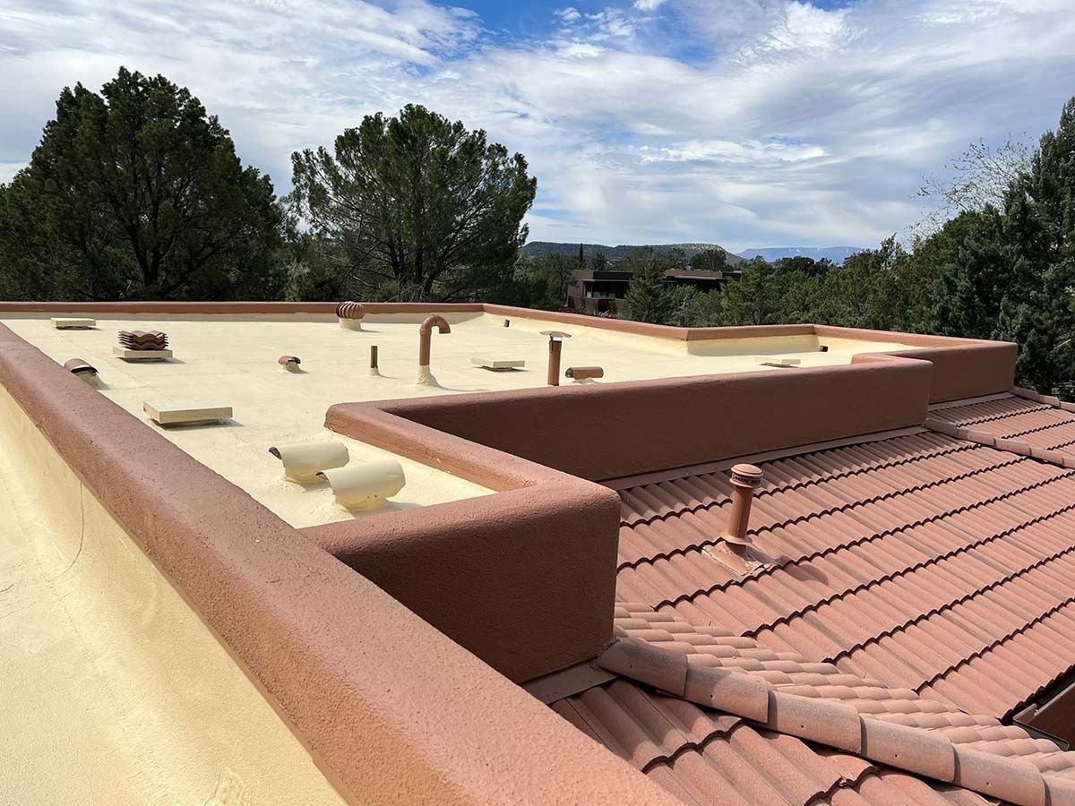 Flat roof with tan coating and terracotta trim, adjacent to a tile roof, under a cloudy sky.