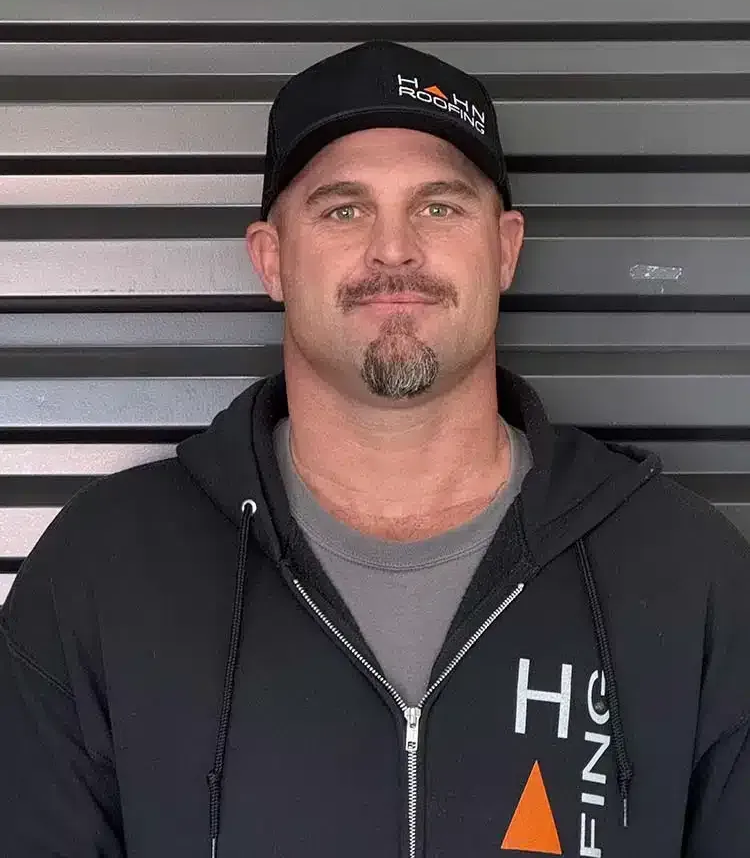 Man in a black hoodie and cap smiles, set against horizontal lines. The cap and hoodie have a logo.