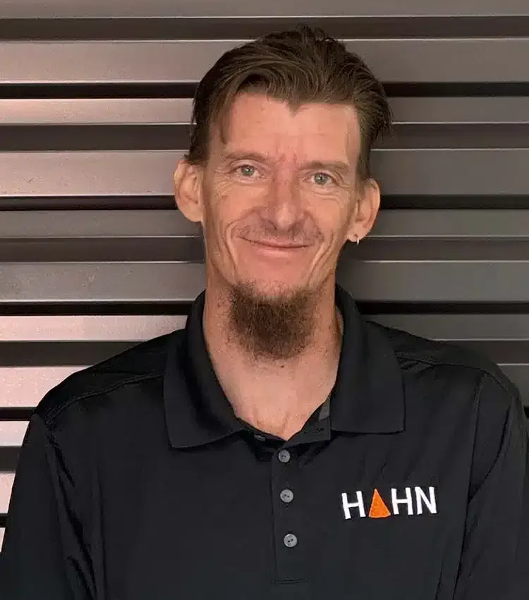 Man with goatee wearing a black polo shirt, smiling in front of a ribbed wall with 