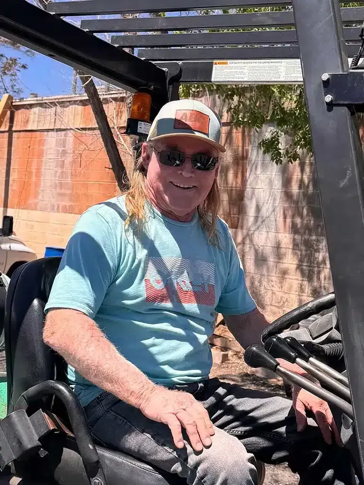 Man in sunglasses and hat driving a forklift outdoors, smiling.
