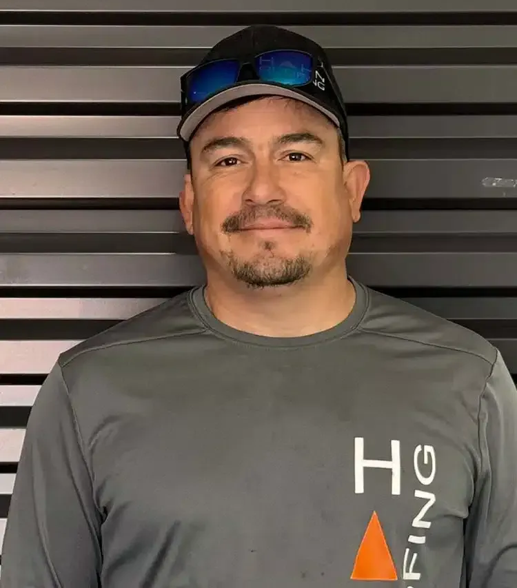 Man wearing a cap and gray shirt, smiling in front of a gray-striped wall. Shirt has an orange triangle and 