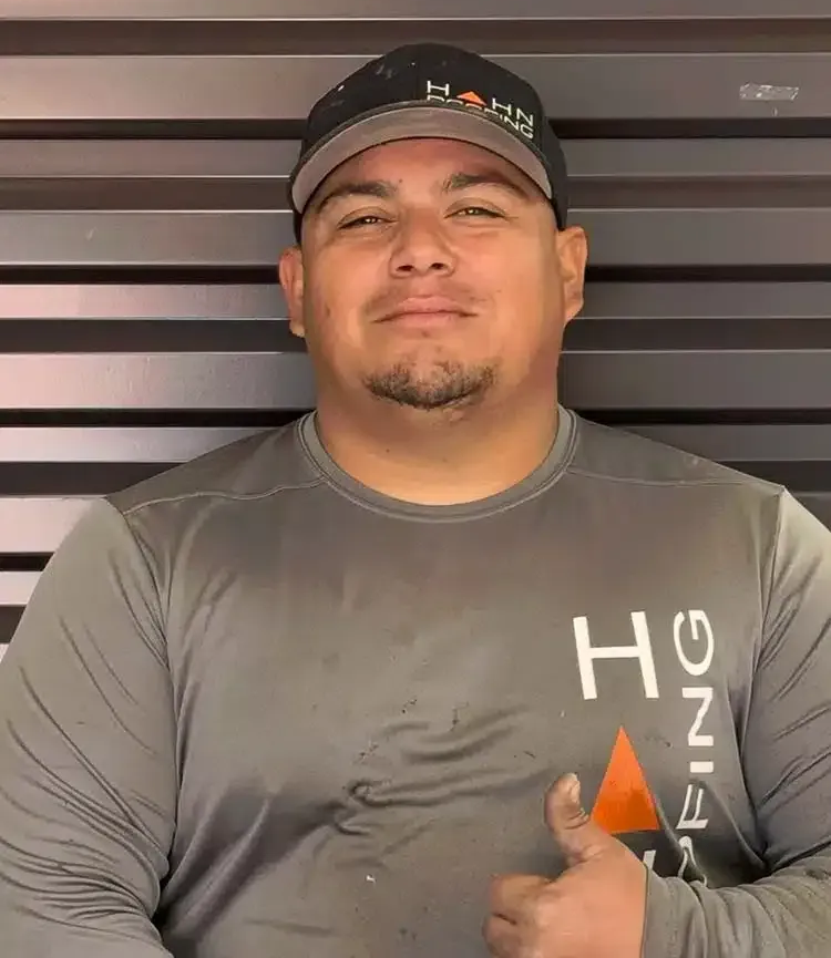 Man in gray shirt and cap with logo, thumbs up, smiles, in front of a metal wall.