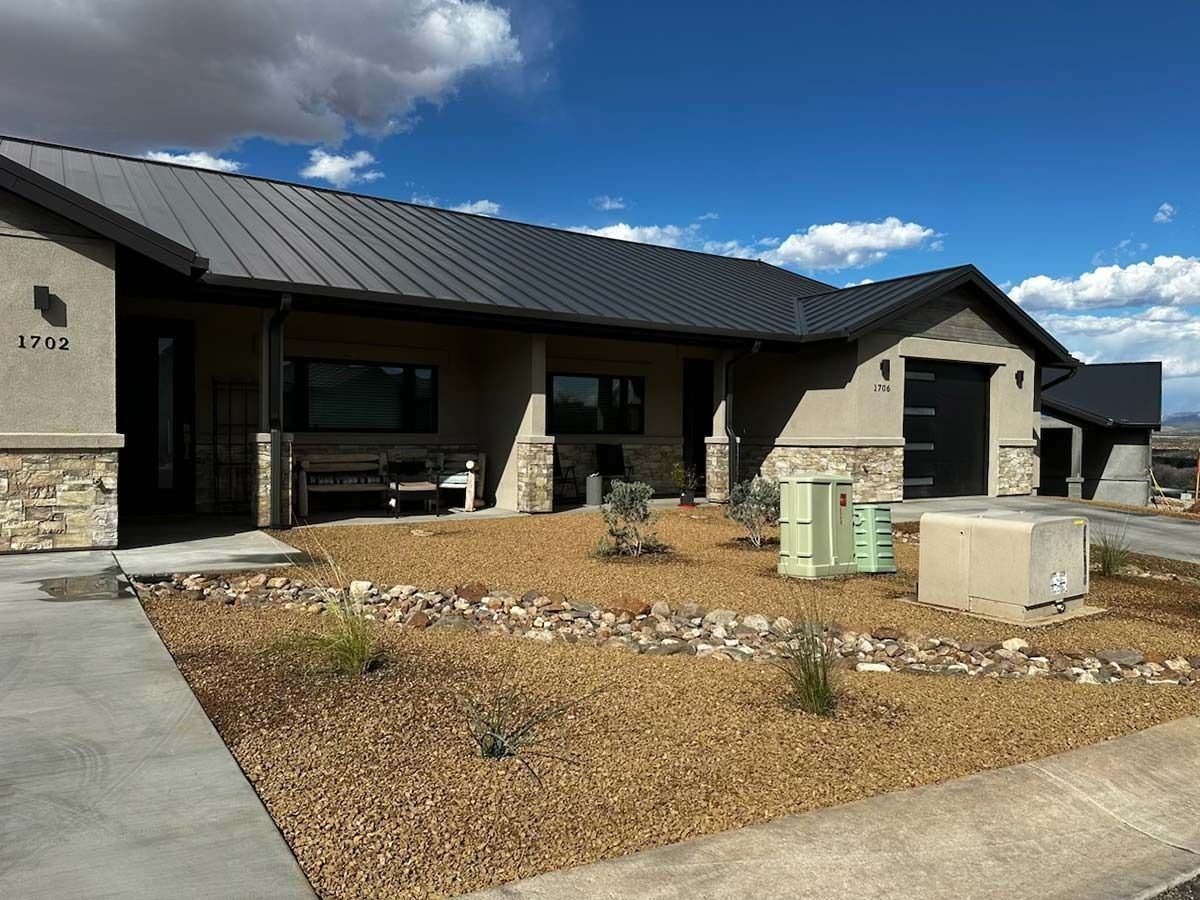Tan and stone house with dark metal roof, gravel landscaping, and blue sky.