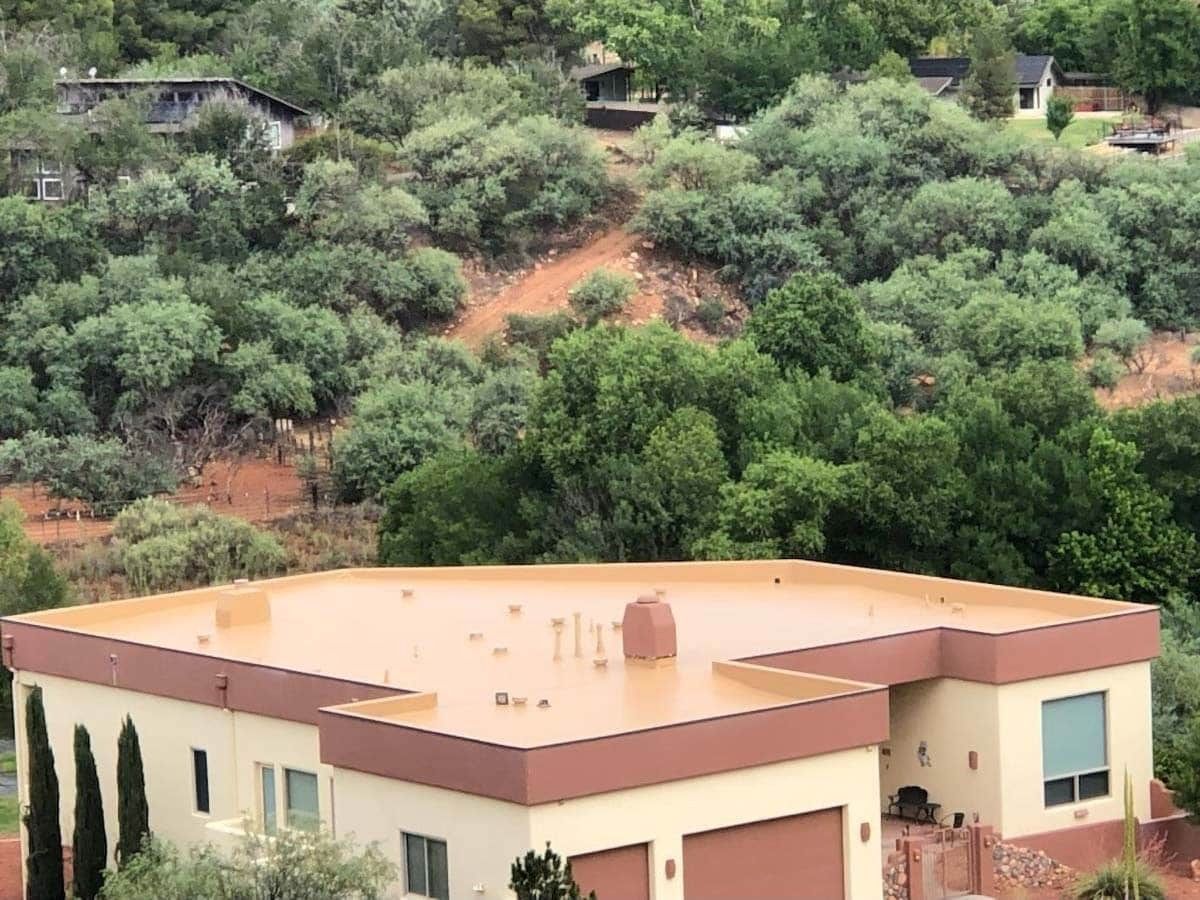 Modern home with flat roof and brown trim against a hillside with green trees and scattered houses.