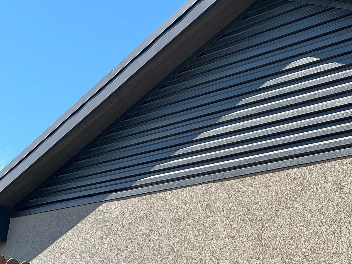 Dark gray slatted vent beneath a roof's overhang, on a stucco wall, against a clear blue sky.