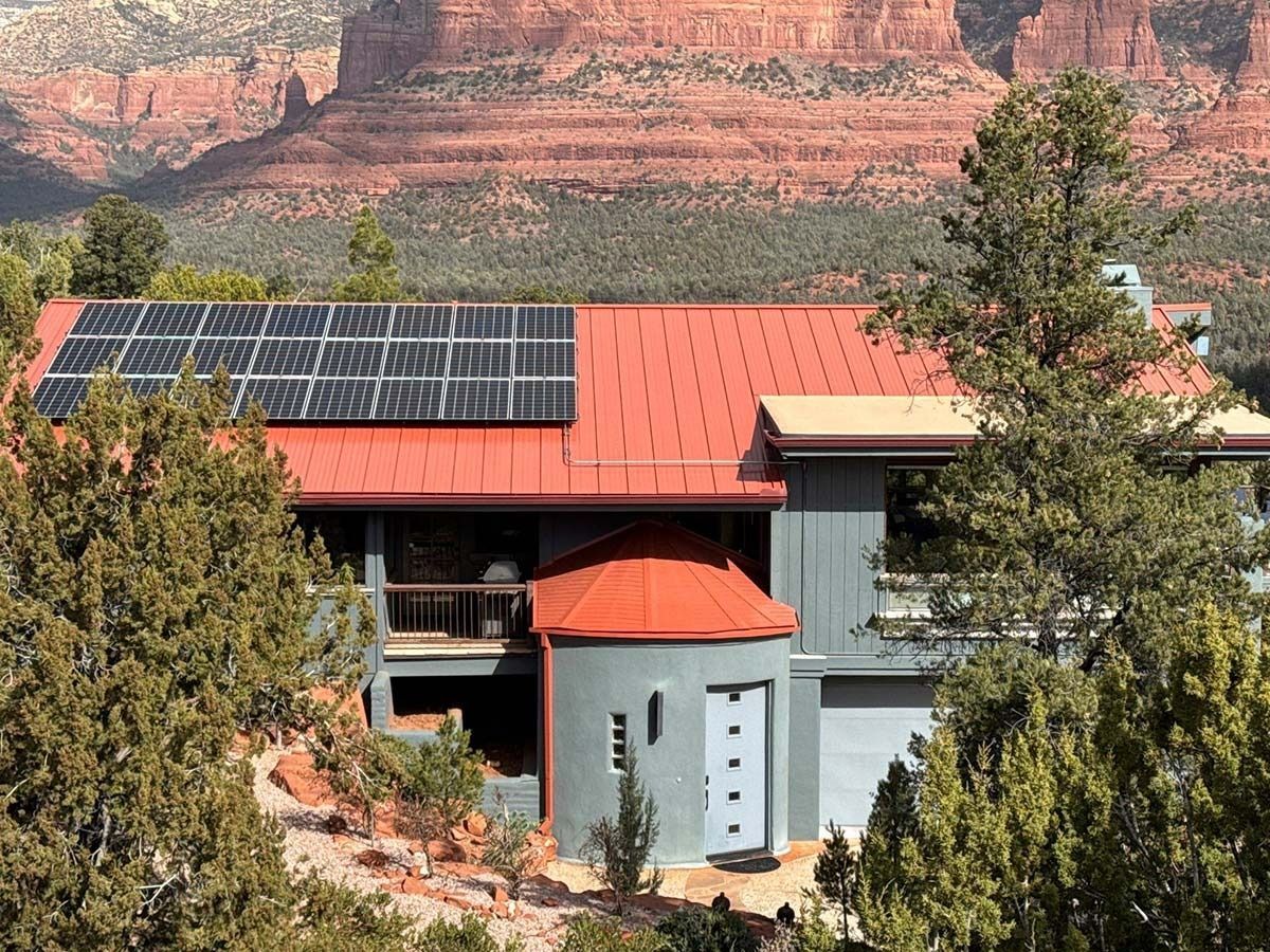 House with red roof, solar panels, and gray siding, set against red rock mountains and green trees.