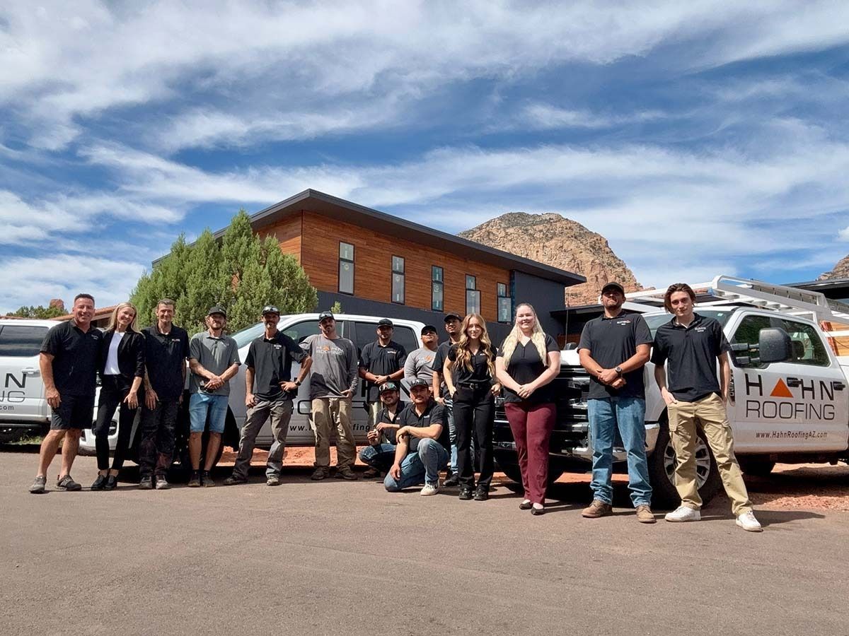 Group of people posing in front of a house, trucks, and a mountain on a sunny day. Some are wearing work clothes.