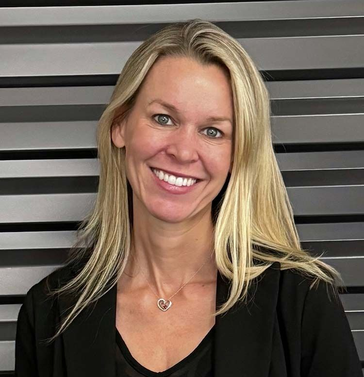 Blonde woman smiles, wearing a black blazer and necklace, against a gray striped background.