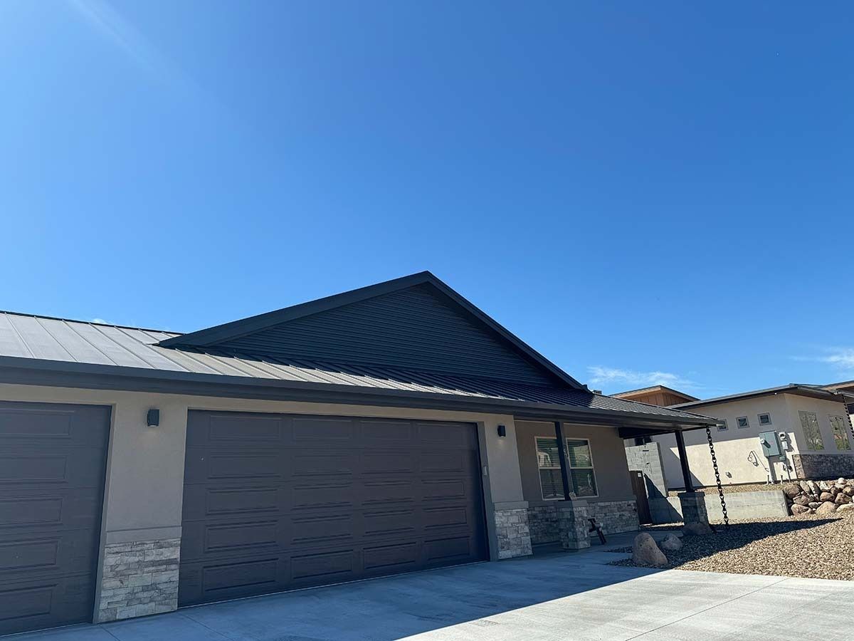 Tan house with dark gray roof and garage doors, stone accents, clear blue sky.