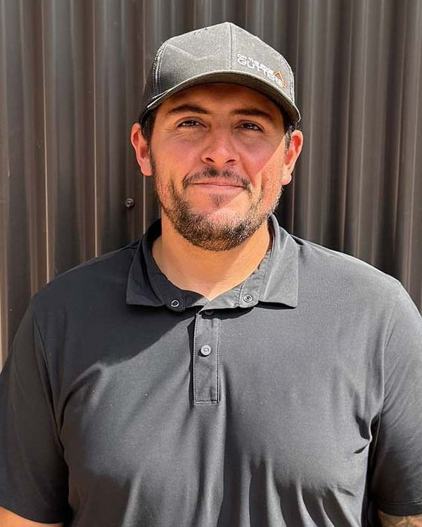 Man in grey polo shirt and cap smiles, standing in front of a dark corrugated wall.