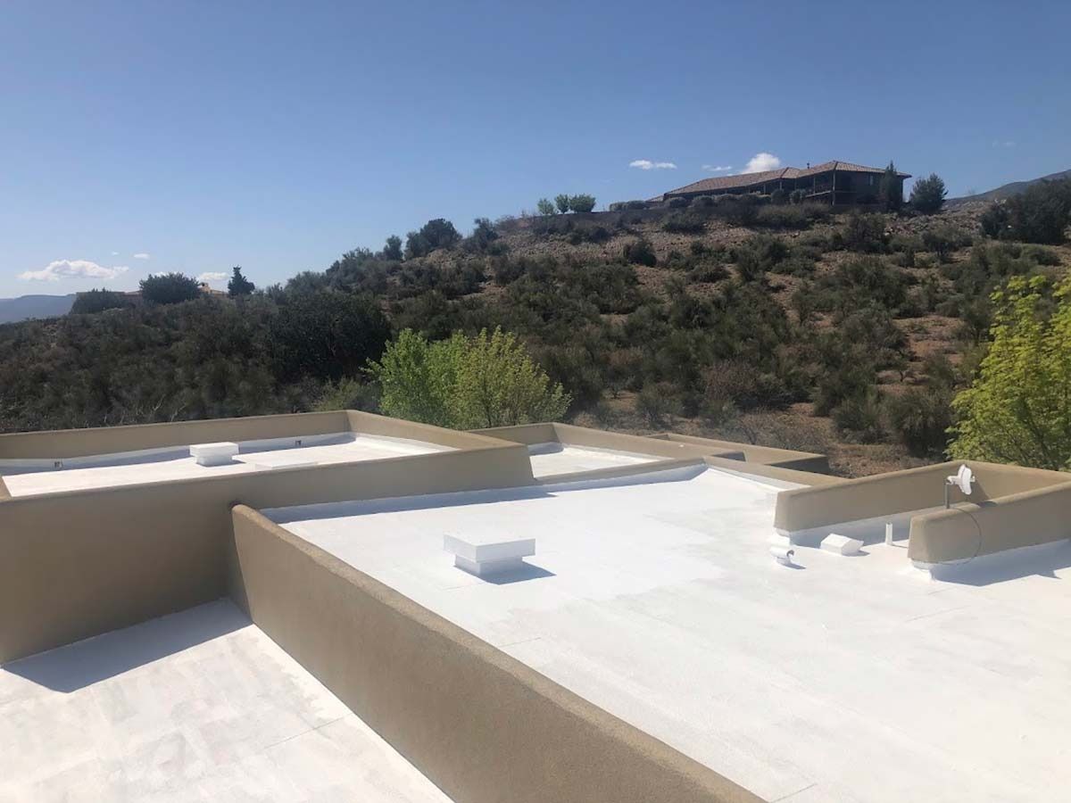 A white flat roof with beige walls. A hillside with vegetation and a building is in the background.
